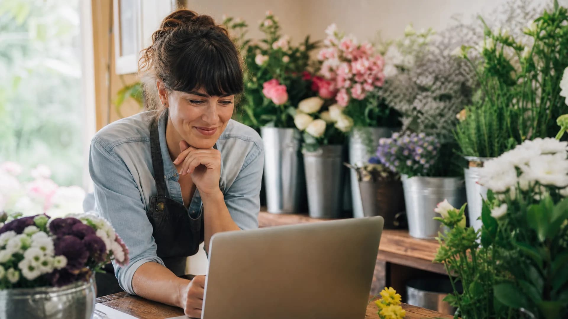 A florist smiling at her laptop, surrounded by fresh flowers in her shop